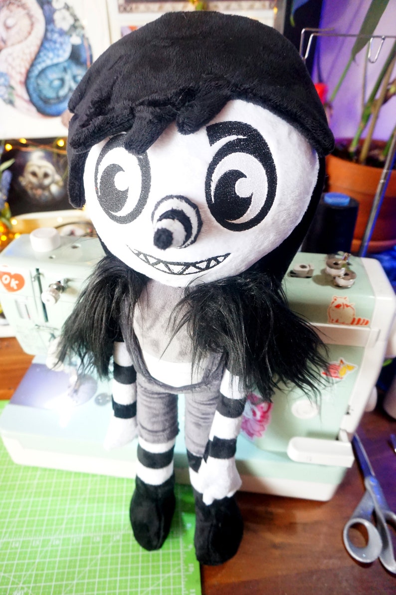 May include: A black and white plush toy with a creepy smile, large eyes, and a long black wig. The toy is wearing a black and white striped outfit and black shoes.