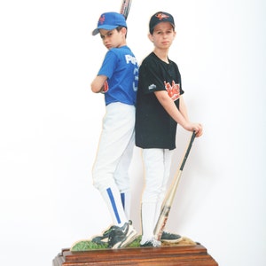 May include: Two young boys wearing baseball uniforms stand side-by-side holding baseball bats. The boy on the left is wearing a blue jersey with the word "Pirates" on it. The boy on the right is wearing a black jersey with the word "Orioles" on it. They are both smiling and looking at the camera.