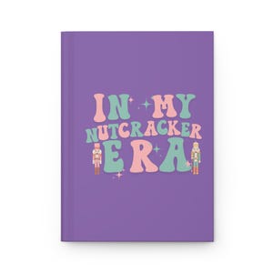 Puede incluir: Un diario morado con el texto "IN MY NUTCRACKER ERA" en letras rosas y turquesas. El diario presenta ilustraciones de cascanueces y estrellas. La portada es de color morado liso.