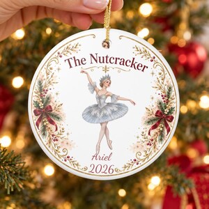 May include: A round white ceramic ornament featuring a ballerina in a blue tutu, surrounded by a gold and red holiday wreath. The text reads The Nutcracker, Ariel, and 2026. It hangs from a gold string against a blurred Christmas tree.