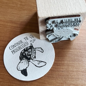 Könnte beinhalten: Ein weißer runder Aufkleber mit einer Schwarzweißillustration einer Schildkröte mit dem Text "CONTINUE, TU PROGRESSES!" in Schwarz.