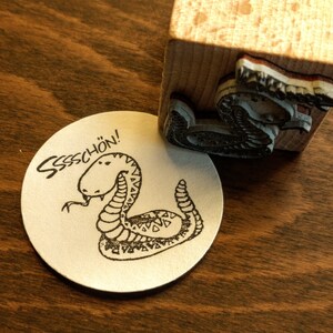 Könnte beinhalten: Ein Gummistempel mit einem Cartoon-Schlangen-Design und dem deutschen Wort "Sssschön!" auf einem weißen, kreisförmigen Papier. Der Stempel ist auf einem Holzblock montiert. Die Schlangenillustration ist in Schwarzweiß gehalten.