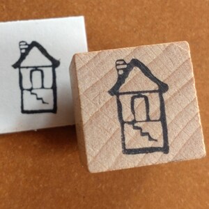 Puede incluir: Un sello de madera con un dibujo de l&iacute;nea negra de una casa con una chimenea y escalones. La casa tiene una puerta y una ventana.