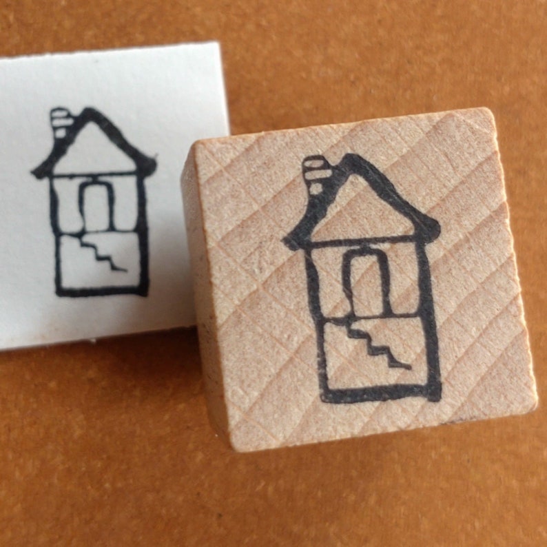 Puede incluir: Un sello de madera con un dibujo de l&iacute;nea negra de una casa con una chimenea y escalones. La casa tiene una puerta y una ventana.