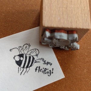 Könnte beinhalten: Ein Holzstempel mit einem Cartoon-Bienen-Design und den Worten "Super fleißig!" auf weißem Papier gedruckt. Die Biene ist schwarz und weiß mit großen Augen und Flügeln. Der Stempel ist teilweise sichtbar.