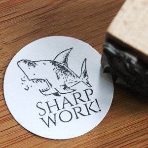 Könnte beinhalten: Ein weißer runder Aufkleber mit einer Schwarzweißillustration eines Hais mit offenem Maul. Der Text "SHARP WORK!" ist unter dem Hai gedruckt.