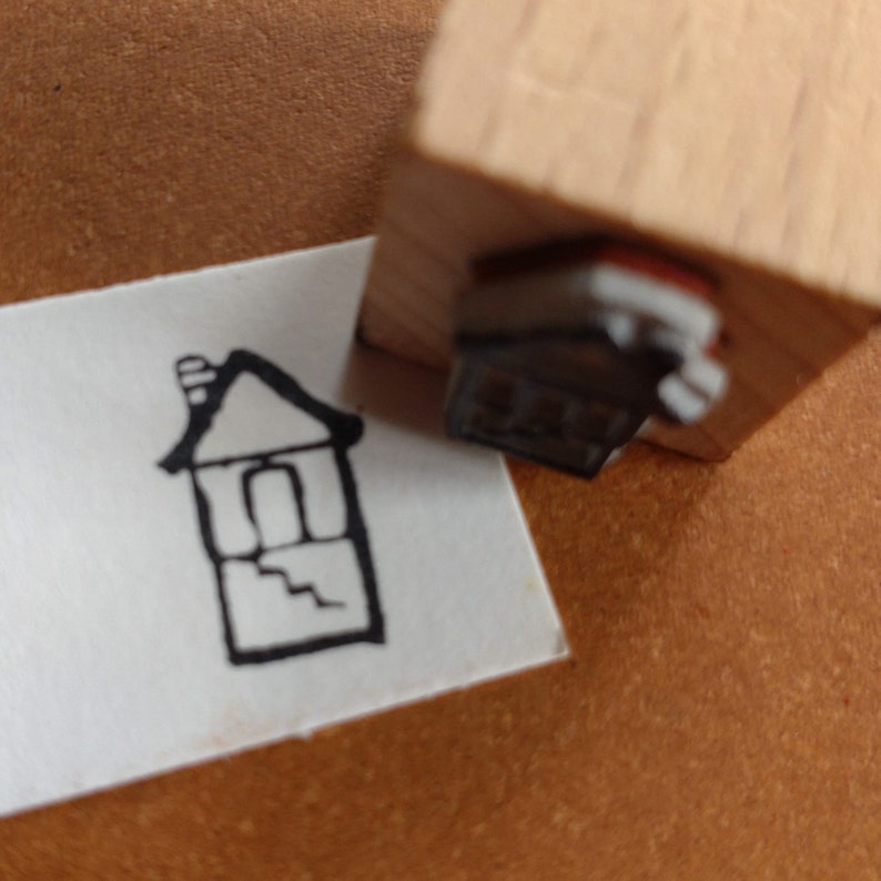 Puede incluir: Una ilustraci&oacute;n en blanco y negro de una casa con una chimenea y una puerta. La ilustraci&oacute;n est&aacute; en una tarjeta blanca y se est&aacute; estampando con un sello de madera.