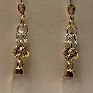 Puede incluir: Pendientes colgantes con un gancho dorado y un adorno en forma de lágrima. Los pendientes presentan anillos plateados, anillos dorados entrelazados y un colgante de piedra blanca en forma de lágrima. Los pendientes son para orejas perforadas.