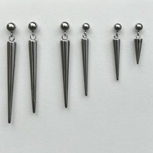 May include: A set of six silver-toned metal earrings arranged in a row on a white display card. Each earring features a small spherical stud top connected to a long, slender, pointed spike pendant that tapers to a sharp tip at the bottom.