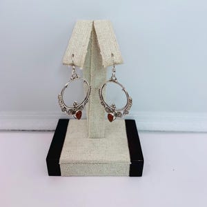 May include: A pair of silver-toned hoop earrings with a decorative design and a small, red gemstone at the bottom. The earrings are displayed on a beige earring stand against a white background.