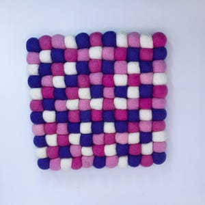 May include: A square trivet constructed from numerous small, round felt balls arranged in a grid pattern. The felt balls feature a repeating color scheme of deep purple, bright magenta, soft pink, and white, creating a textured surface.