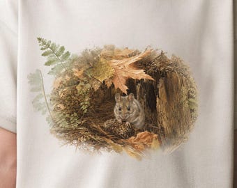 May include: A light beige short-sleeve t-shirt featuring a central graphic print of a small brown mouse nestled inside a hollow tree trunk surrounded by autumn leaves, pinecones, and a green fern frond in a detailed, realistic artistic style.