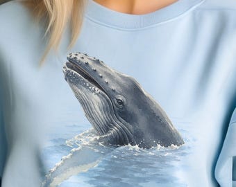 May include: A light blue crewneck sweatshirt featuring a detailed graphic illustration of a humpback whale breaching the surface of the water. The whale is depicted in shades of dark gray and white, showing its head and pectoral fin above the waves.