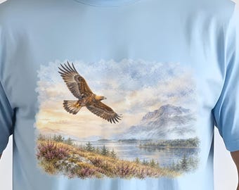 May include: A light blue t-shirt featuring a graphic print of a golden eagle soaring above a landscape with a lake, mountains, and purple heather. The artwork is rendered in a painterly style with soft clouds and natural tones across the center chest.