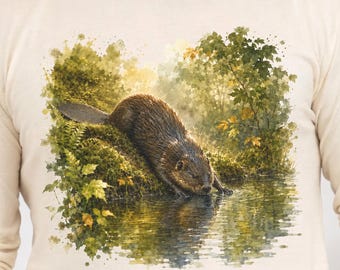 May include: A cream-colored long-sleeve shirt featuring a detailed watercolor-style graphic of a brown beaver resting on a mossy, green riverbank. The illustration depicts the beaver near the water's edge, surrounded by lush green foliage.