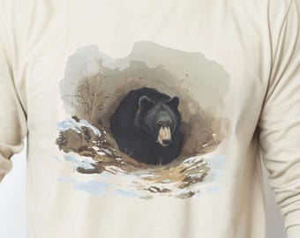 May include: A long-sleeve crew neck shirt in a light beige color featuring a graphic print on the chest. The print depicts a black bear resting inside a den surrounded by patches of snow and dry grass, rendered in a painterly, illustrative style.