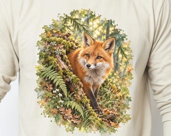 May include: A long-sleeve beige crewneck shirt featuring a detailed graphic print of a red fox emerging from a mossy, fern-covered forest log. The illustration uses vibrant green, brown, and orange tones to depict the fox in its natural habitat.