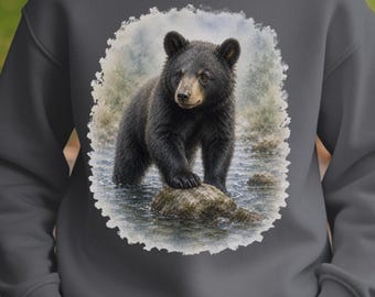 May include: A charcoal gray crewneck sweatshirt featuring a central graphic print of a black bear standing on a rock in a shallow stream. The illustration is framed by a soft, textured white border, contrasting with the solid dark gray fabric.