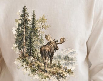 May include: A cream-colored crewneck sweatshirt featuring a detailed graphic print of a moose standing in a forest landscape with tall pine trees, a lake, and distant mountains. The illustration uses earthy tones of brown, green, and muted gray.