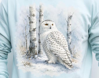 May include: A light blue long-sleeve sweatshirt featuring a detailed watercolor-style graphic of a white snowy owl perched in a winter forest with birch trees and a soft, snowy ground. The design is centered on the chest of the garment.