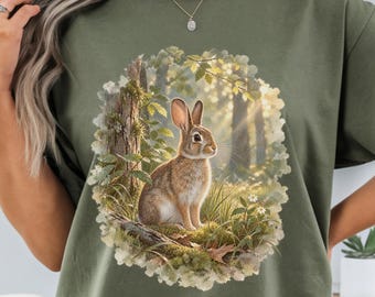 May include: An olive green short-sleeve t-shirt featuring a circular graphic print of a brown rabbit sitting in a sunlit forest clearing surrounded by green foliage, mossy tree trunks, and small white flowers, with a crew neckline and loose fit.