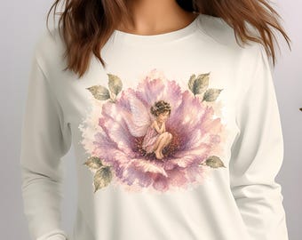 May include: A long-sleeve cream-colored shirt featuring a central graphic of a small fairy with wings sitting inside a large, watercolor-style pink and purple flower with green leaves. The design is positioned on the chest of the garment.