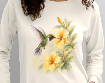 May include: A long-sleeve cream-colored shirt featuring a graphic print of a hummingbird hovering near yellow hibiscus flowers. The bird has iridescent green and purple feathers, and the floral design includes green leaves and soft yellow petals.