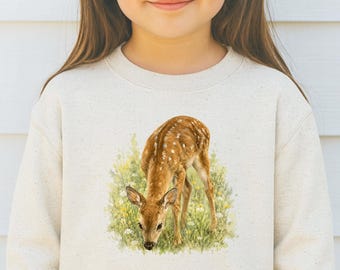 May include: A long-sleeve crewneck sweatshirt in a light oatmeal speckled color. The front features a detailed, realistic illustration of a young spotted fawn standing in a patch of green grass and yellow wildflowers, rendered in soft, natural tones.