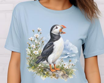 May include: A light blue t-shirt featuring a graphic illustration of a puffin standing on a rock among white flowers with a coastal background. The puffin has black and white feathers, an orange beak, and orange feet, depicted in a watercolor style.