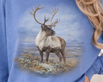 May include: A periwinkle blue long-sleeve crewneck sweatshirt featuring a detailed graphic print of a reindeer standing in a tundra landscape. The reindeer has large antlers and a thick coat, depicted in shades of brown, gray, and white.