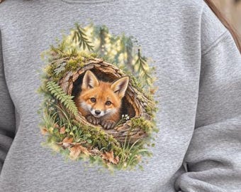 May include: A heather gray long-sleeve sweatshirt featuring a central graphic print of a small orange fox peeking out from a hollow log surrounded by green moss, ferns, and foliage. The garment has a crew neckline and ribbed cuffs and hem.