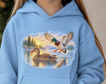 May include: A light blue hooded sweatshirt featuring a graphic print of two mallard ducks on a lake. One duck is swimming in the water while the other is landing with wings spread. The design includes a forest background and a sunrise sky.