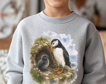 May include: A light gray crewneck sweatshirt featuring a graphic illustration of an adult puffin standing on a rocky cliff next to a small puffin chick nestled in a burrow. The design includes soft watercolor textures with blue sky and sea.