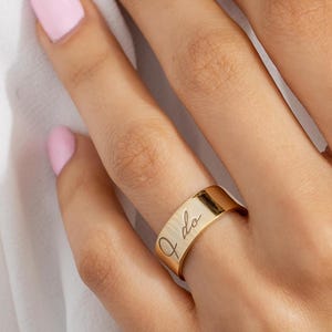 以下が含まれることがあります： 「I do」の文字が繊細な筆記体で刻まれたゴールドカラーのリング。リングは幅広のバンドスタイルで、光を反射します。ピンクのマニキュアを塗った指にリングがはめられています。