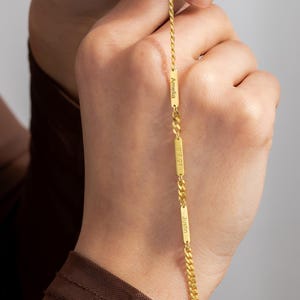 May include: A gold-colored necklace with a delicate chain and rectangular bar pendants. Each bar pendant has a name engraved on it. The necklace is held up against a neutral background.