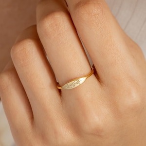 May include: A gold ring with a leaf design. The ring is worn on a finger, and the design is centered on the ring's face. The ring is set against a white background.