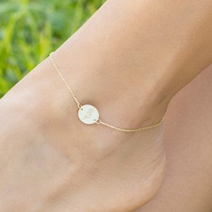 May include: A delicate gold anklet with a small round charm featuring a floral design. The anklet is shown on a person's ankle.
