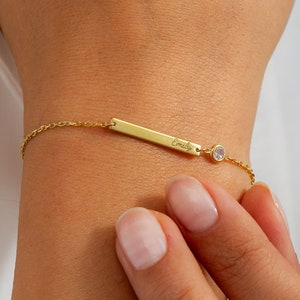 Op de afbeelding: Gouden armband met een kleine, heldere stenen bedel en de naam "Emily" gegraveerd op de staaf.
