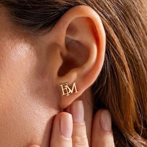 May include: Close-up of a person's ear with a pair of gold stud earrings. The earrings feature the letters "EM" in a serif font. The person's hand is gently touching their face near the ear. The earrings are a simple, elegant design.