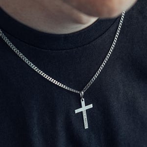 May include: A silver cross pendant necklace with a curb chain. The cross has engraved text on the vertical bar. The necklace is worn against a dark background.