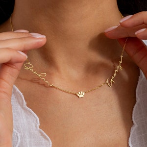 May include: Gold necklace with two names, "Emilia" and "Henry", written in cursive script. A small paw print charm hangs between the names.