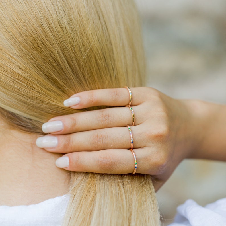 May include: Four thin rose gold rings, each featuring a series of small, multicolored gemstones set along the band. The rings are worn on four fingers, showcasing a delicate design with vibrant, tiny stones in various hues including blue, green, and yellow.