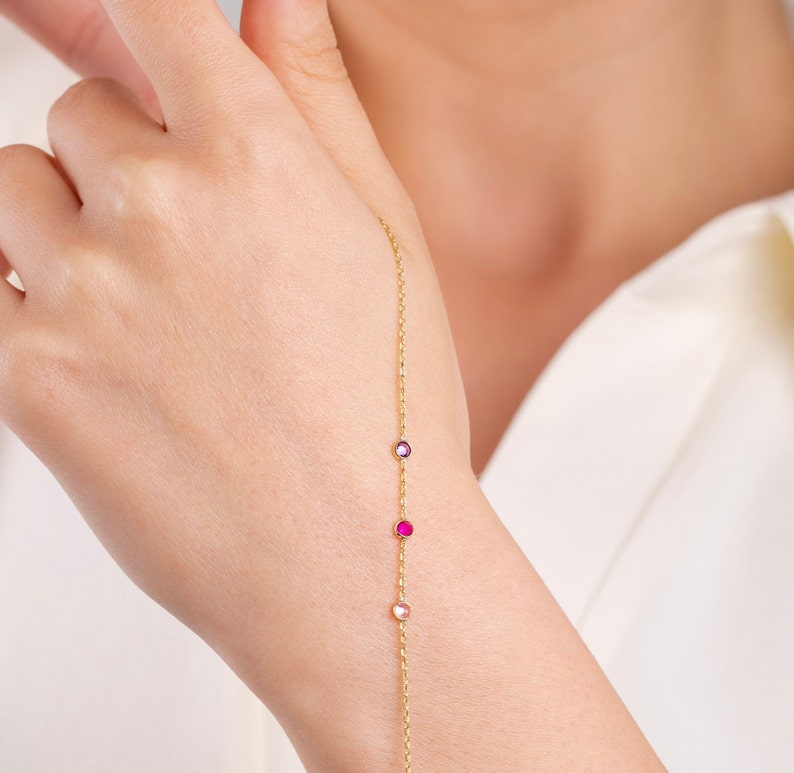 May include: A delicate gold chain bracelet with three small gemstones. The gemstones are purple, pink, and clear.