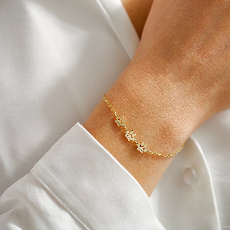 May include: A gold chain bracelet with three paw print charms. Each charm has a small inscription.