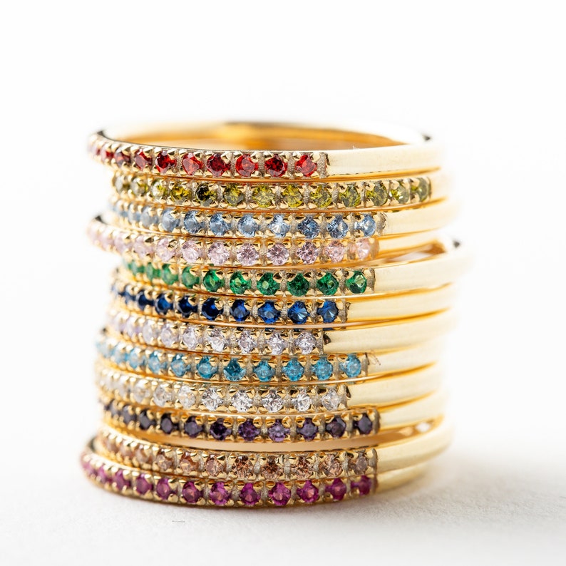 May include: A stack of gold rings with a single row of colorful gemstones around each band. The rings are stacked in a rainbow order, starting with red and ending with purple.