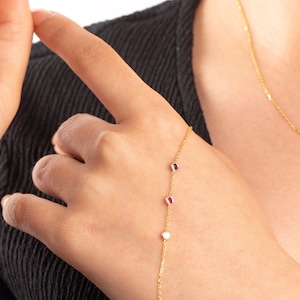 May include: A gold chain bracelet with three small oval-shaped gemstones. The gemstones are red, white, and purple.