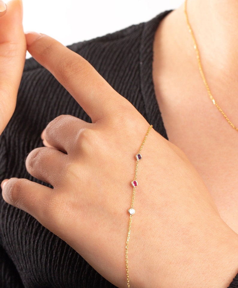 May include: A gold chain bracelet with three small oval-shaped gemstones. The gemstones are red, white, and purple.