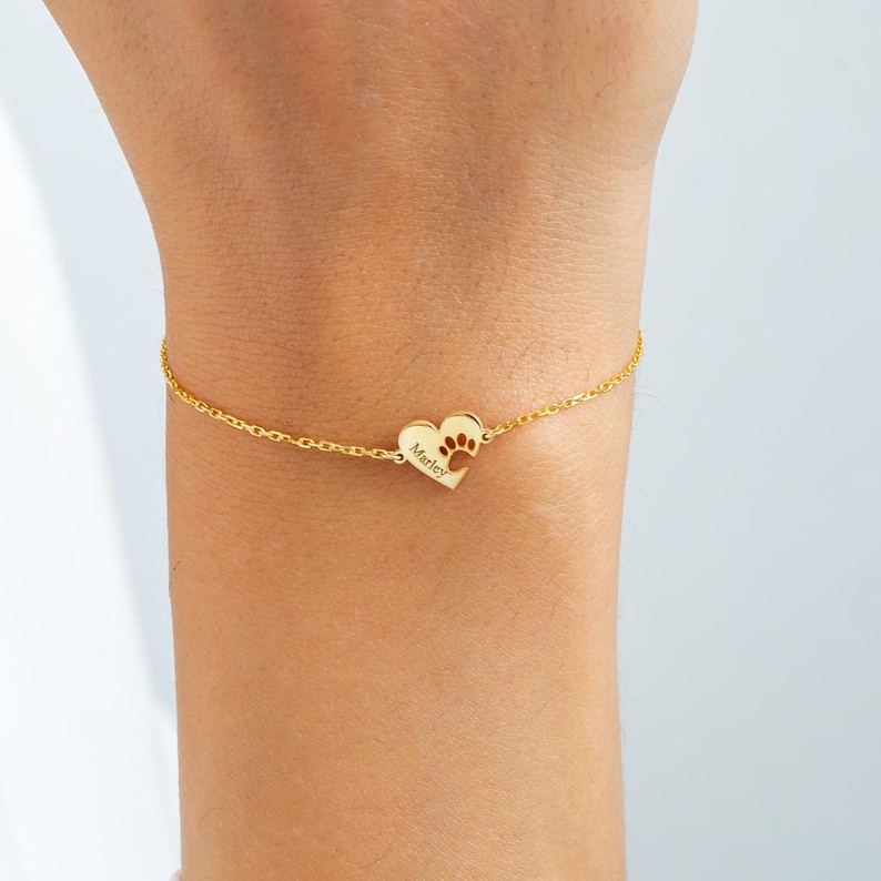 May include: Gold-coloured bracelet with a heart-shaped charm. The charm has a paw print and the name "Marley" engraved. The bracelet is delicate and made of a fine chain.