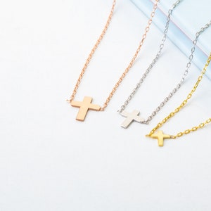 Pu&ograve; includere: Tre collane con croce in oro rosa, argento e oro. Ogni collana presenta un semplice pendente a forma di croce sospeso a una catena sottile. Le collane sono esposte su una superficie bianca e azzurra.