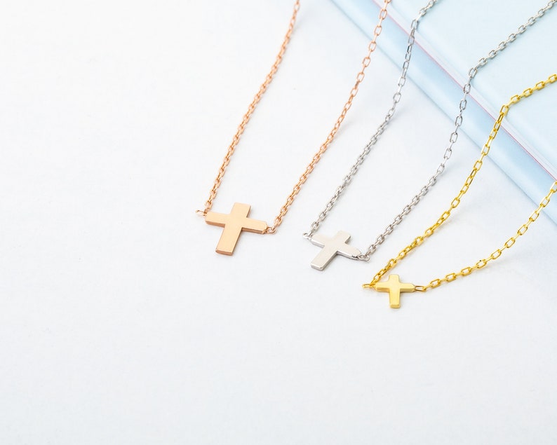 Pu&ograve; includere: Tre collane con croce in oro rosa, argento e oro. Ogni collana presenta un semplice pendente a forma di croce sospeso a una catena sottile. Le collane sono esposte su una superficie bianca e azzurra.